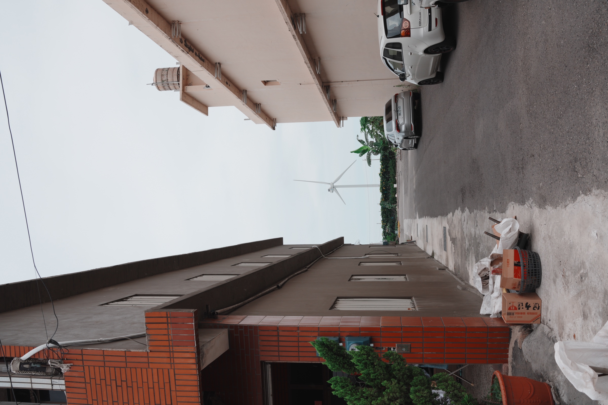 A wind turbine seen at the end of a residential lane in Hanbao, between houses, parked cars, and utility wires